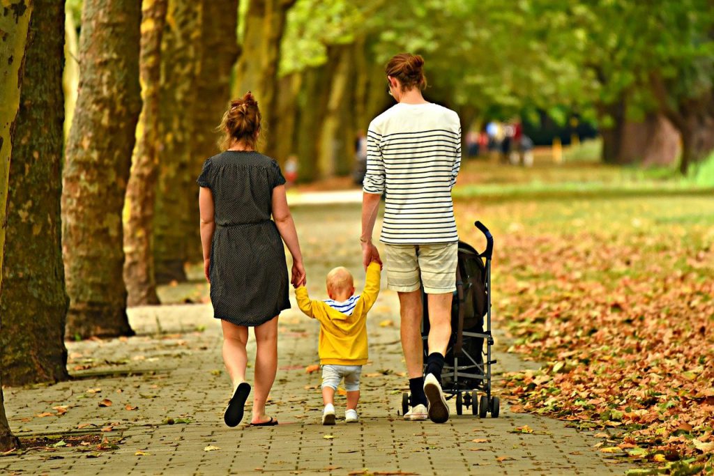 woman wearing a dark dress, child wearing a yellow hoodie, man wearing shorts and a shirt pushing a stroller down a path with leaves on ground in a park
