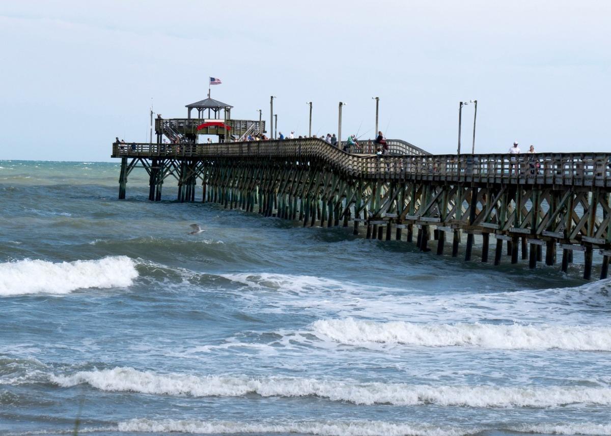 pier viewed from land at a slight angle with people lined up along rail fishing, rough ocean